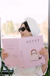 Person in a white robe with a towel turban and black sunglasses, smiling while reading a pink luxury fashion coffee-table book featuring a handbag illustration, seated by a bright window with houseplants.