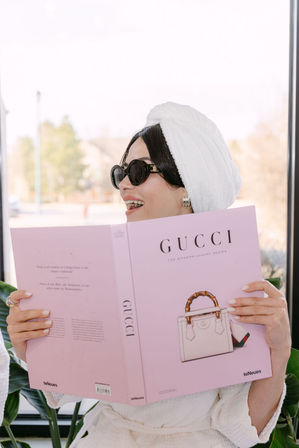 Person in a white robe with a towel turban and black sunglasses, smiling while reading a pink luxury fashion coffee-table book featuring a handbag illustration, seated by a bright window with houseplants.