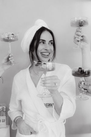 Black-and-white photo of a smiling woman in a spa robe and towel turban raising a champagne coupe in a celebratory toast with friends.