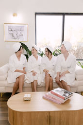 Four friends in white spa robes and towel turbans laughing on a cream sofa in a bright modern living room, enjoying an at-home spa day with plants, natural light, a wooden coffee table and stacked fashion books.