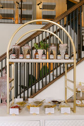 Gold-arched display shelf with amber dropper bottles, decorative glass jars and labeled bowls of dried herbs (rose bud, calendula, lemongrass, jasmine buds) and a small potted plant, staged in front of a wooden staircase.