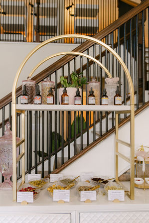 Gold-arched display shelf with amber dropper bottles, decorative glass jars and labeled bowls of dried herbs (rose bud, calendula, lemongrass, jasmine buds) and a small potted plant, staged in front of a wooden staircase.