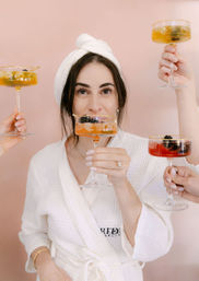 Spa-day celebration: woman in a white robe and towel turban toasts with friends' colorful coupe cocktails against a blush pink backdrop