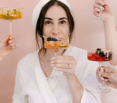 Spa-day celebration: woman in a white robe and towel turban toasts with friends' colorful coupe cocktails against a blush pink backdrop