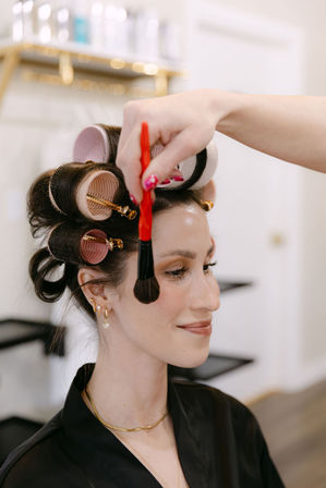 Smiling woman in a beauty salon with pink and white hair rollers as a stylist uses a red-handled makeup brush to apply blush to her cheek.