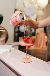 Hand pouring a pink canned beverage into a pink coupe glass filled with ice and cherries — bright cocktail moment at a stylish party bar