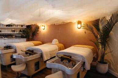 Cozy boutique spa pedicure room with three plush treatment chairs, white linens and towels, warm wall sconces, airy fabric ceiling drapes and potted palms for a relaxing vibe.