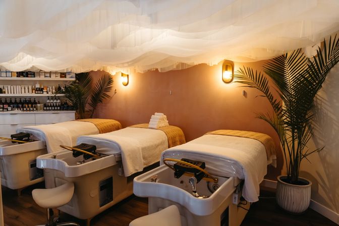 Cozy boutique spa pedicure room with three plush treatment chairs, white linens and towels, warm wall sconces, airy fabric ceiling drapes and potted palms for a relaxing vibe.