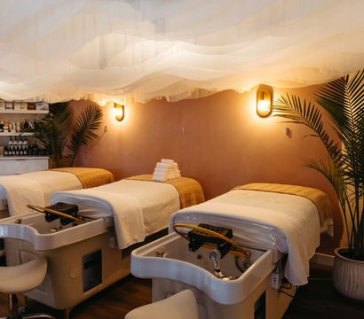 Cozy boutique spa pedicure room with three plush treatment chairs, white linens and towels, warm wall sconces, airy fabric ceiling drapes and potted palms for a relaxing vibe.