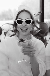 Black-and-white photo of a smiling woman in a spa robe with towel turban and cat-eye sunglasses, holding a fruit-topped cocktail glass.
