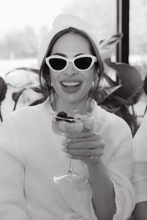 Black-and-white photo of a smiling woman in a spa robe with towel turban and cat-eye sunglasses, holding a fruit-topped cocktail glass.