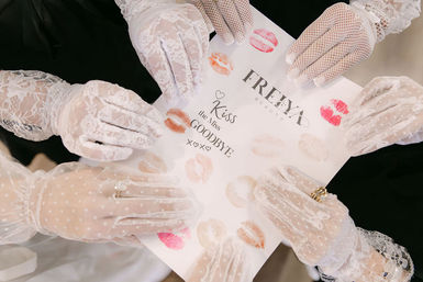 Overhead view of multiple hands in white lace gloves holding a sheet stamped with pink, red and nude lipstick kiss prints — playful vintage-style bridal party keepsake.