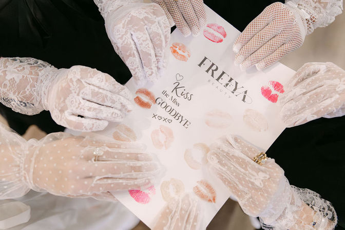 Overhead view of multiple hands in white lace gloves holding a sheet stamped with pink, red and nude lipstick kiss prints — playful vintage-style bridal party keepsake.