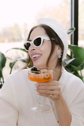 Chic spa-day scene: smiling woman in a white robe and towel headwrap wearing white sunglasses and sparkly earrings, holding a coupe with an orange cocktail garnished with lychee and a blackberry against an indoor plant backdrop.
