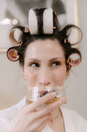 Close-up of a woman with pink hair rollers and gold clips sipping a drink while getting ready — playful beauty prep and hair-styling moment.