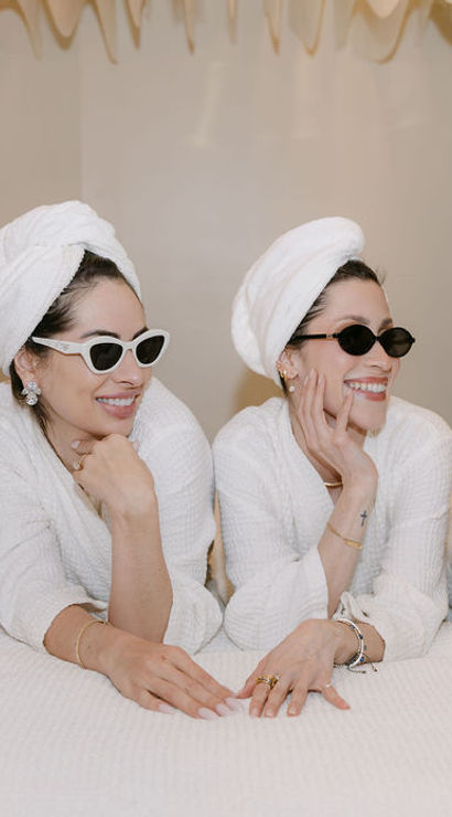 Four women enjoying a spa day in white robes and towel turbans, wearing sunglasses and laughing together while lounging on treatment beds in a bright indoor spa lounge