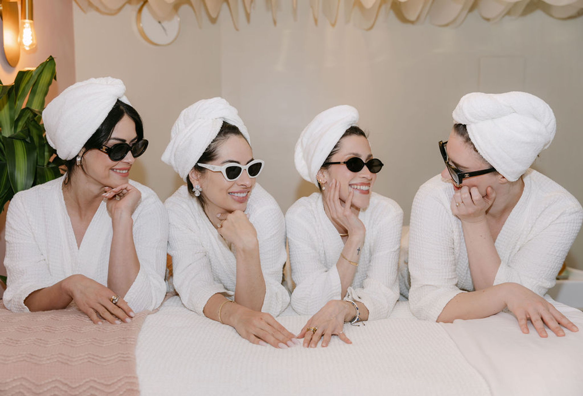 Four women enjoying a spa day in white robes and towel turbans, wearing sunglasses and laughing together while lounging on treatment beds in a bright indoor spa lounge