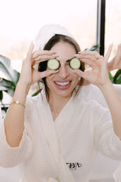 Woman in white robe and towel holds cucumber slices over her eyes, smiling during a relaxing at-home spa facial in a bright indoor setting