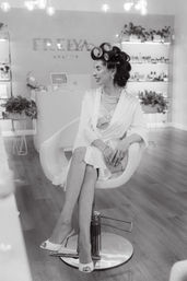 Black-and-white photo of a woman in a robe with hair rollers, under-eye patches and pearl necklace sitting in a modern beauty salon chair, smiling while prepping for a special occasion.