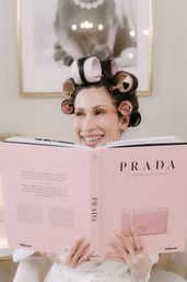 Smiling woman with pink hair rollers reading a large pink fashion book in a bright modern room