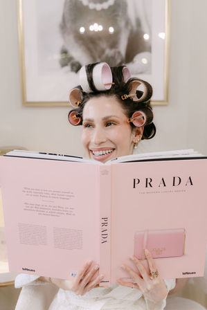 Smiling woman with pink hair rollers reading a large pink fashion book in a bright modern room