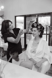Hairstylist sprays hairspray on a smiling client with large hair rollers in a bright beauty salon, the seated woman wearing a robe, lace camisole and pearl necklace.