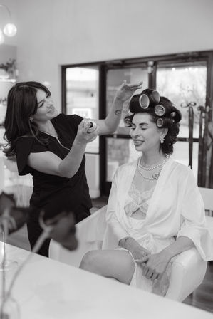 Hairstylist sprays hairspray on a smiling client with large hair rollers in a bright beauty salon, the seated woman wearing a robe, lace camisole and pearl necklace.