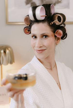 Smiling woman in a white robe with pink hair rollers and gold clips, holding a champagne coupe with berries in a cozy spa-style pampering moment.