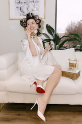 Retro-glam woman in a white satin robe with hair rollers and pearls lounges on a white sofa in a bright living room, smiling while holding a gold vintage telephone; white high heels with red soles and an ornate gold rotary phone nearby.