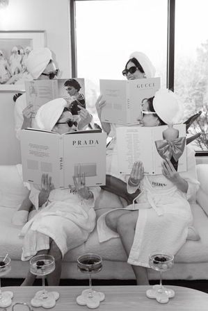 Four friends in white spa robes and towel turbans wearing sunglasses, lounging on a bright living room sofa and reading oversized fashion coffee-table books, with champagne coupes on the coffee table — playful at-home spa and style gathering.