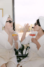Two women in white spa robes and towel turbans at a sunny window, wearing sunglasses and toasting colorful cocktails while feeding each other blackberry garnishes