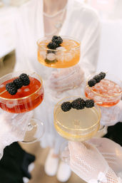 Four gold-rim coupe cocktails in red, amber and pale yellow, topped with blackberry skewers and ice, held by hands in white lace gloves at an elegant gathering