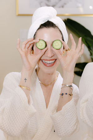 Playful woman in a white robe and towel turban holding cucumber slices over her eyes and smiling during an at-home spa facial and skincare routine.