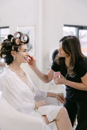 Woman in white robe and pearl necklace with hair rollers having lipstick applied by a makeup artist in a bright beauty salon — bridal makeup and hair prep