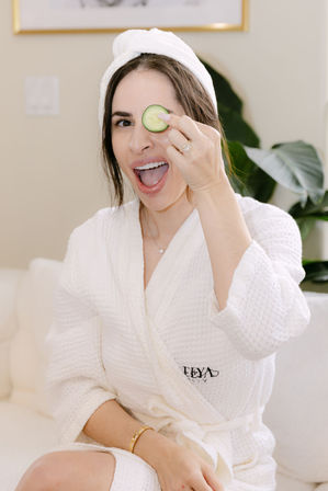 Playful woman in a white waffle robe and towel turban holding a cucumber slice over one eye, smiling during an at-home spa facial and skincare pampering session.