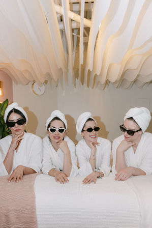 Four women in white spa robes and towel turbans wearing sunglasses, lounging on a treatment bed beneath a wavy fabric ceiling in a chic boutique spa, playful spa day vibe.