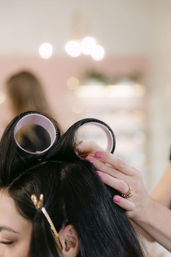 Close-up of a stylist placing large velcro rollers into long dark hair in a bright beauty salon, showing hands with pink nails, a gold ring and a hair clip.
