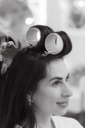 Black-and-white portrait of a smiling woman at a beauty salon with large hair rollers clipped to her crown while a stylist adjusts them.