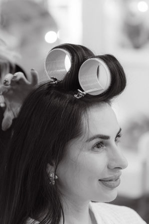 Black-and-white portrait of a smiling woman at a beauty salon with large hair rollers clipped to her crown while a stylist adjusts them.