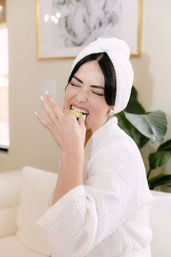 Person in a white bathrobe with a towel turban playfully biting a cucumber slice during an at-home spa moment on a bright couch with a leafy plant and framed art in the background.