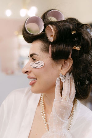 Woman in a beauty salon with pink hair rollers and under-eye patches, wearing a pearl necklace and lace glove while adjusting a sparkly earring