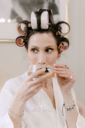 Close-up of a woman in a white robe with pink hair rollers and gold clips sipping champagne from a coupe glass during a get-ready moment