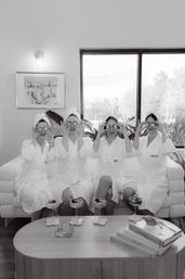 Black-and-white image of four women in white spa robes and towels on a couch holding cucumber slices over their eyes during a cozy at-home spa day with cocktails on the coffee table and stacked fashion books.
