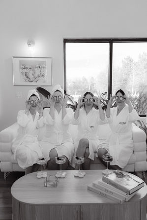 Black-and-white image of four women in white spa robes and towels on a couch holding cucumber slices over their eyes during a cozy at-home spa day with cocktails on the coffee table and stacked fashion books.