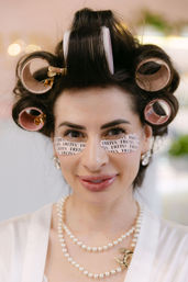 Close-up of a smiling woman in vintage-glam hair rollers and gold clips, printed under-eye patches under her eyes, wearing pearl necklace and earrings during beauty prep.