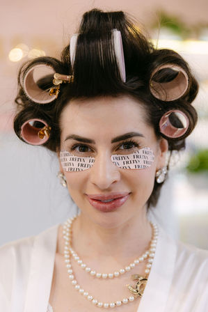 Close-up of a smiling woman in vintage-glam hair rollers and gold clips, printed under-eye patches under her eyes, wearing pearl necklace and earrings during beauty prep.