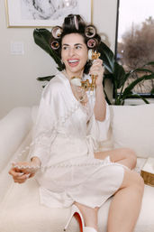 Smiling woman in a white satin robe with hair rollers and pearl necklace laughing while holding a vintage gold telephone handset on a cream sofa in a stylish at-home getting-ready scene