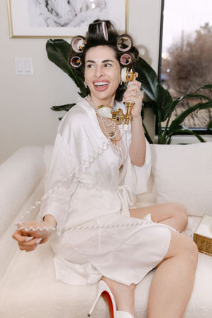 Smiling woman in a white satin robe with hair rollers and pearl necklace laughing while holding a vintage gold telephone handset on a cream sofa in a stylish at-home getting-ready scene