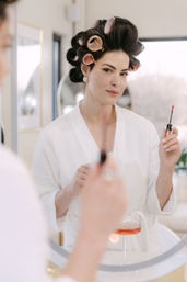 Woman in white robe with pink hair rollers applying lip gloss at a round vanity mirror in a bright beauty salon, relaxed getting-ready makeup routine with a cocktail on the counter