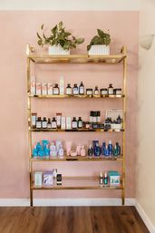 Gold metal shelf against a blush-pink wall displaying assorted skincare and haircare bottles, jars and pastel packaging with two potted trailing plants on top — chic retail beauty display.